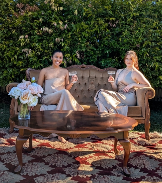 Two women seated on a vintage carved settee with a wooden coffee table in a garden setting
